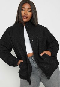 Czarna, teksturowana kurtka z guzikami z przodu, w oversize'owym kroju i kieszeniami, w połączeniu z białą bluzką i szarymi dżinsami.