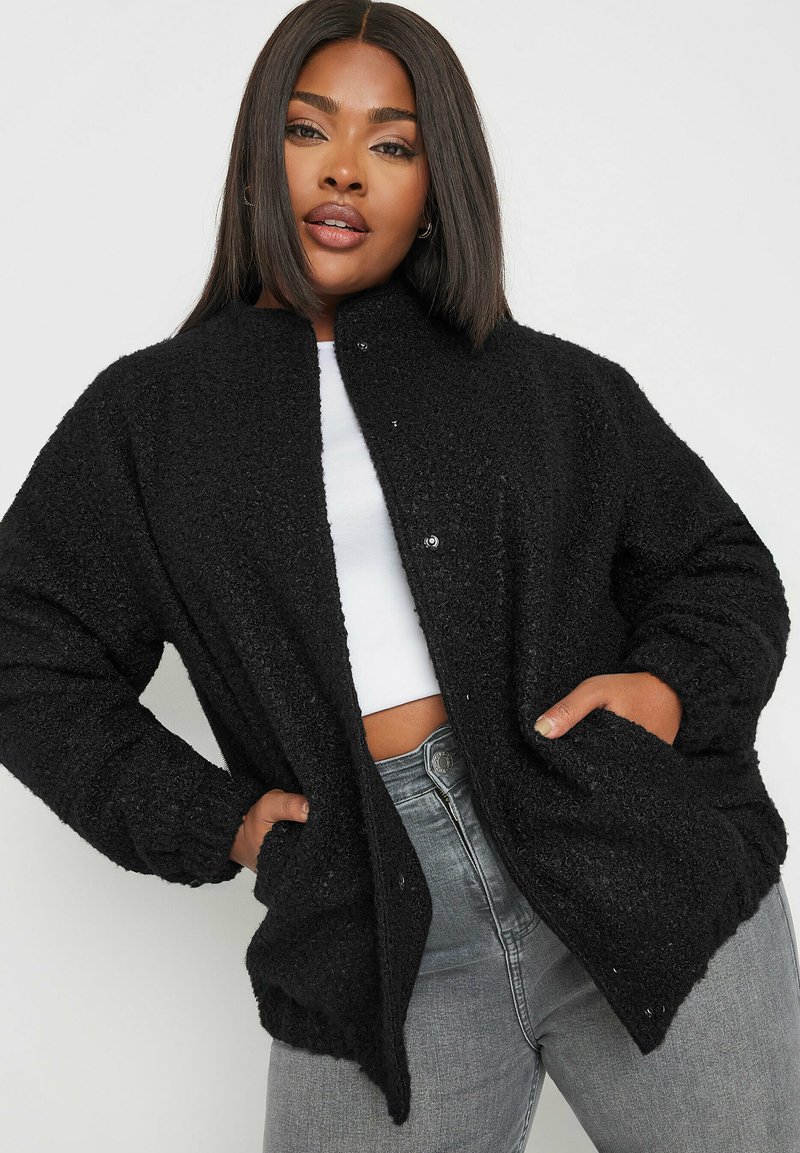 Czarna, teksturowana kurtka z guzikami z przodu, w oversize'owym kroju i kieszeniami, w połączeniu z białą bluzką i szarymi dżinsami.