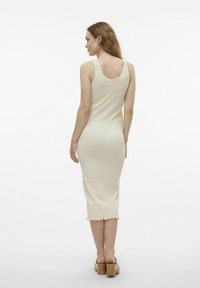 Robe tricotée beige, longueur genou, à côtes avec de larges bretelles et un ourlet en forme de vagues. Texture lisse, silhouette ajustée, portée avec des sandales à talons épais.