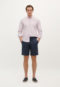Cămașă roz deschis cu dungi, cu nasturi și mâneci lungi, asociată cu pantaloni scurți bleu navy și pantofi de barcă bleu navy. Design simplu, fără modele vizibile.