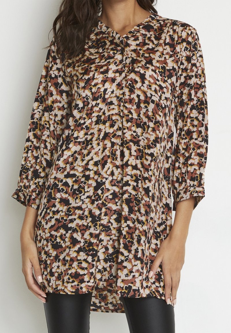 Femme portant un chemisier ample à manches longues avec un motif floral abstrait multicolore et un pantalon noir ajusté.