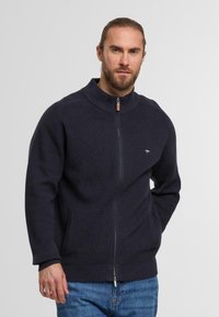 Marineblauer Strickzipper mit hohem Kragen, geripptem Muster und seitlichen Taschen. Mit kleinem Logo auf der Brust und einem glatten Reißverschluss.