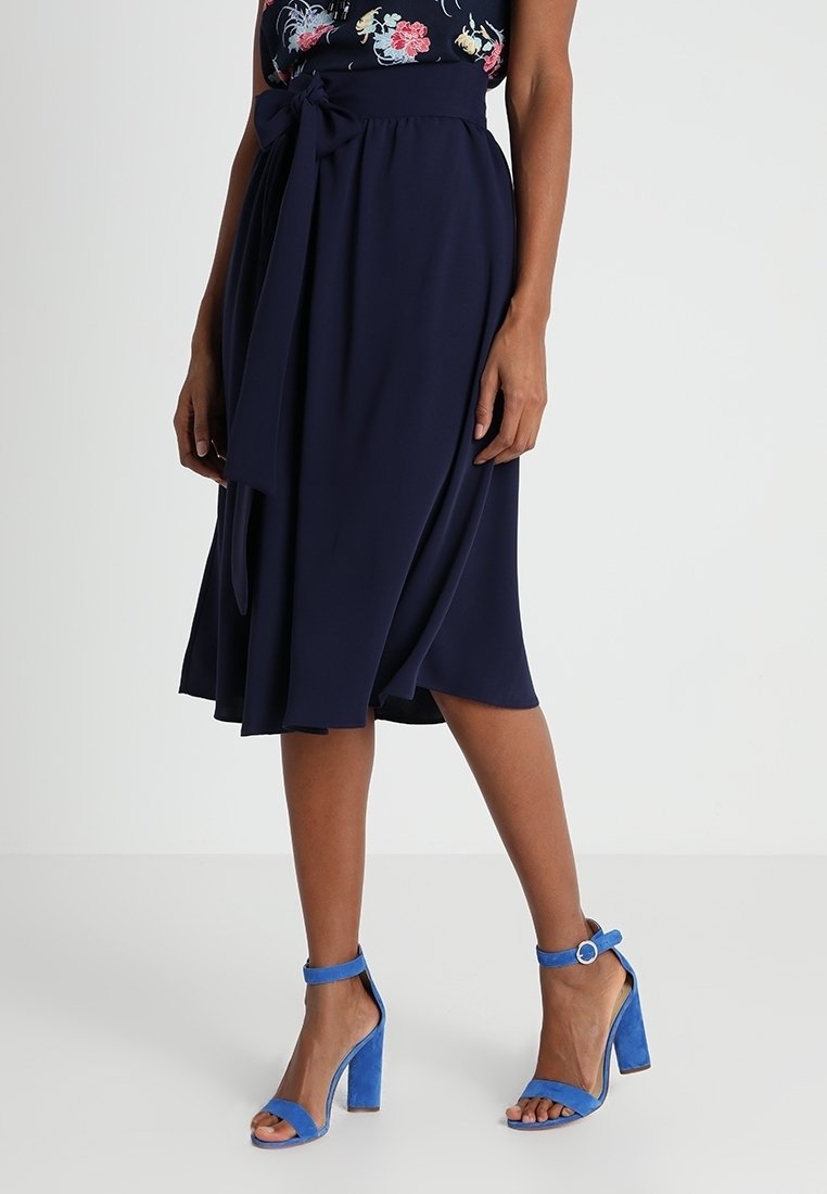Femme portant une jupe midi bleu marine avec une ceinture nouée à la taille, associée à des sandales à talons hauts bleu vif à bouts ouverts avec des brides à la cheville.