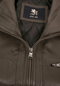 Braune Lederjacke mit Reißverschluss vorne, gestepptem Innenfutter und genähten Details. Innen sichtbar ist ein Etikett mit einem Logo.