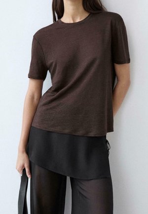 Personne portant un t-shirt marron à manches courtes sur une jupe asymétrique noire et un pantalon noir transparent, tenant une sangle noire dans une main.