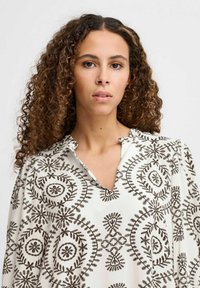 Langærmet bluse i hvidt stof med detaljeret brun broderi med cirkulære og blomster mønstre, samt en krave med indhak.
