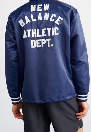 Person iført en marineblå jakke med "NEW BALANCE ATHLETIC DEPT." i hvide bogstaver på ryggen, kombineret med mørke shorts.