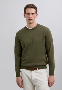 Zielono-oliwkowy dzianinowy sweter z okrągłym dekoltem, ściągaczami na rękawach i u dołu, zestawiony z białymi spodniami oraz brązowym plecionym paskiem.