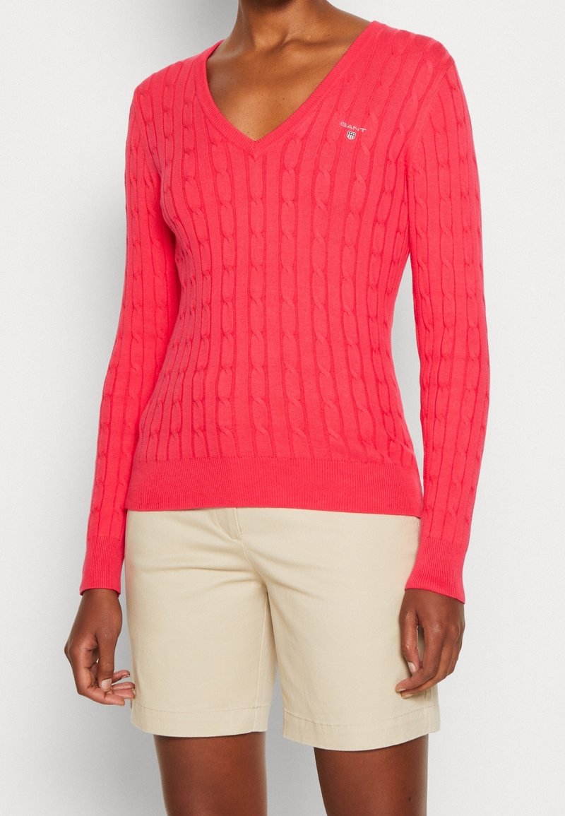 Femme portant un pull en maille torsadée corail vif à col en V et un short beige mi-long, debout sur fond blanc.