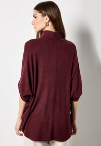 Suéter burdeos de gran tamaño con cuello de embudo, mangas cortas y un acabado texturizado. Corte holgado con un detalle de costura central en la parte posterior.