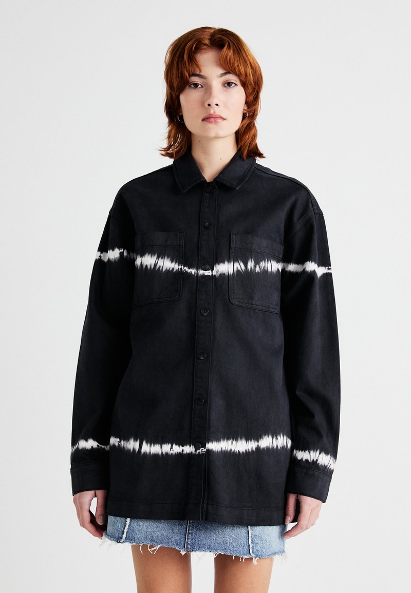Cămașă din denim negru cu un model alb tie-dye în mijloc, cu un guler butonat și două buzunare pe piept. Design lejer, oversized.