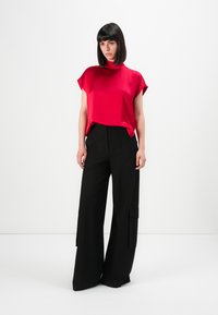 Femme se tenant contre un fond blanc, portant un haut rouge vif à manches courtes et un pantalon noir large avec des poches latérales.