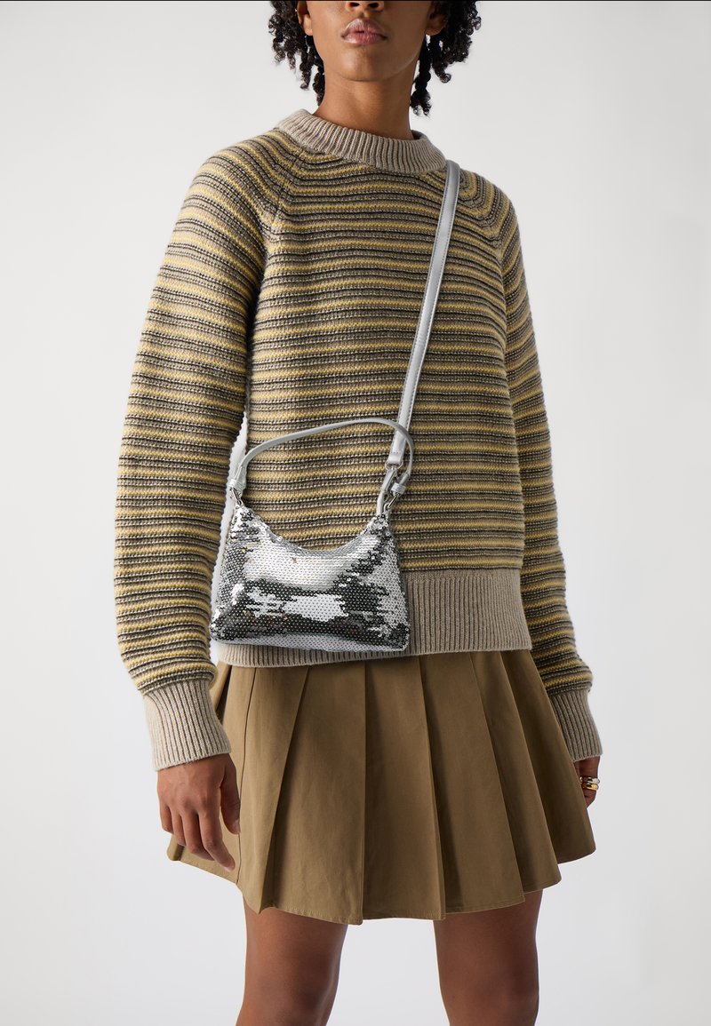 Silver sequined handbag with a curved shape, gray strap, worn over a striped knit sweater and pleated khaki skirt.