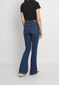 Uppfläkta jeans i mörkblått, med hög midja och bakfickor med kontrasterande sömmar, i kombination med en svart t-shirt och mönstrade skor.