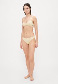 Gaiši dzeltena bikini komplekts, kas ietver trīsstūra krūšturi ar plānām siksnām un atbilstošus zemu vidukļa apakšbikses, abas rotātas ar mazu zilu logo.