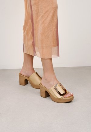 Mules de plataforma color beige claro con un grueso tacón de madera y una amplia hebilla decorativa en un tono más oscuro. Diseño de punta abierta, textura suave.