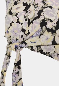 Blommig blus i svart tyg med lila och gula blommor, knappstängning och detalj med knuten knytning vid fållen.