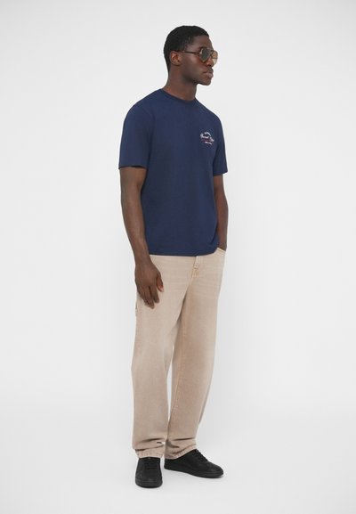 T-shirt en coton marine à manches courtes, arborant un petit motif graphique ; assorti à un pantalon beige à jambes larges et des baskets noires.