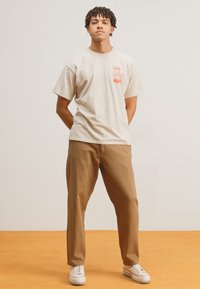 Beige cotton t-shirt with red graphic detail, paired with light brown trousers and white shoes. Model stands with hands behind back.