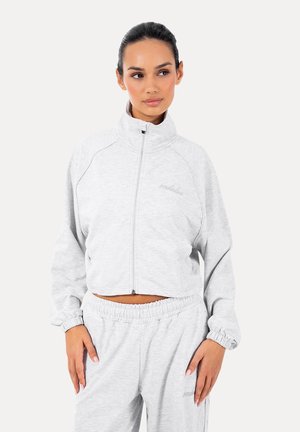 Kobieta z włosami związanymi do tyłu, ubrana w jasnoszarą kurtkę na zamek i pasujące spodnie z elastycznym paskiem, stojąca na jednolitym tle.