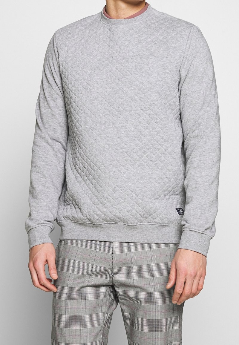 Sweat-shirt matelassé gris avec un col rond et des poignets côtelés, présentant un motif texturé, associé à un pantalon à carreaux.