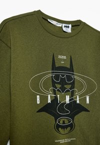 Sudadera verde oliva con gráfico de Batman. Presenta un diseño en blanco y negro con texto. Cuello redondo, tejido suave y estilo minimalista.