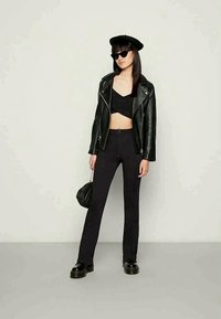 Mulher com jaqueta de couro preta, top curto, calças de cintura alta, sapatos robustos, boina preta, óculos de sol, segurando uma pequena bolsa preta em frente a um fundo simples.