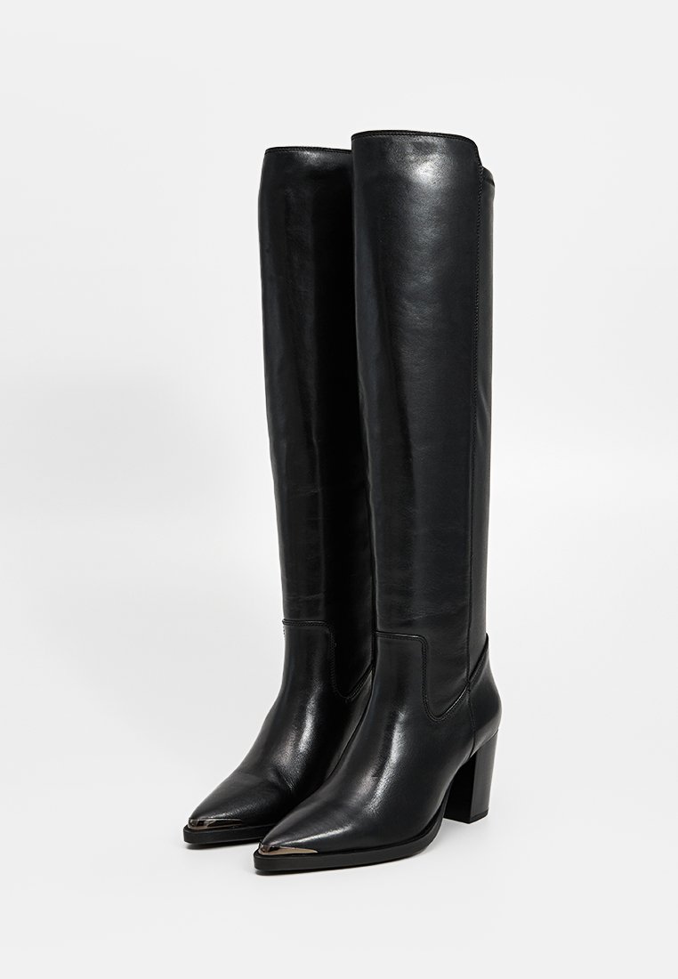 Bottes en cuir noires, montantes jusqu'au genou, avec des bouts pointus, des embouts en métal argenté et des talons hauts épais, présentées debout sur un fond blanc.