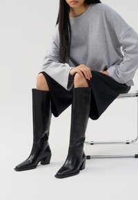 Stivali neri in pelle al ginocchio con punta quadrata e tacco a blocco, caratterizzati da una texture liscia e un design minimalista.