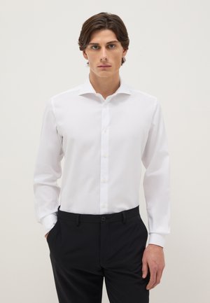 Jeune homme portant une chemise blanche à manches longues boutonnée et un pantalon noir, debout les mains dans les poches devant un fond uni.