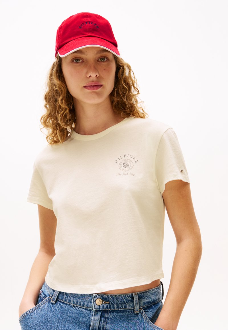 Hvid cropped t-shirt med et lille grafisk tryk i beige, parret med blå højtaljede denimjeans. Modellen bærer en rød cap med hvide detaljer.