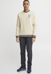 Sudadera color crema con puños y cuello a rayas negras, que presenta un emblema negro de la letra 'P'. Unos pantalones grises y unas zapatillas deportivas azules completan el look.