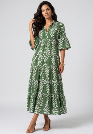 Mujer con cabello largo y oscuro, vistiendo un vestido maxi verde con botones, estampado de hojas blancas y mangas con volantes, posando con una mano tocándose el cabello.