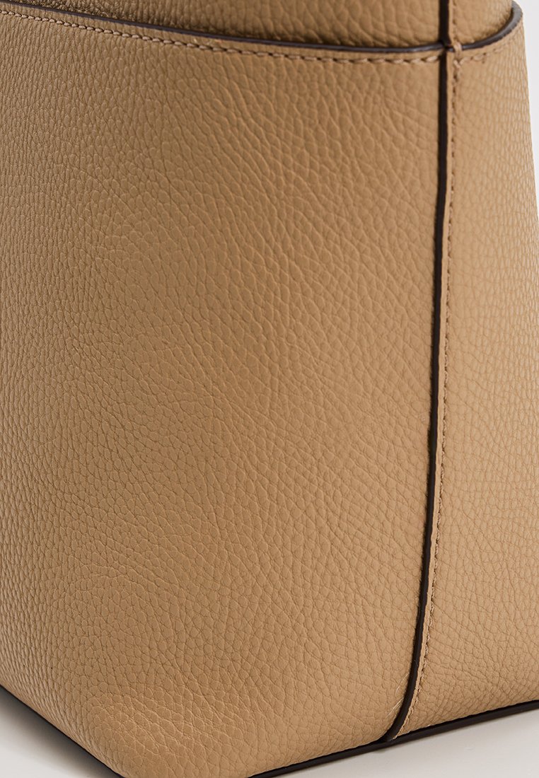 Primo piano di un angolo di una borsa in pelle beige con texture che mostra una fine grana, cuciture e bordi lisci su uno sfondo neutro.