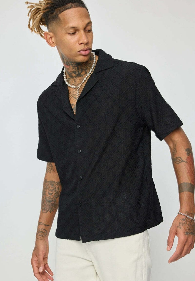 Chemise noire à manches courtes avec un motif texturé, col et fermeture à boutons, portée sur un pantalon de couleur claire.