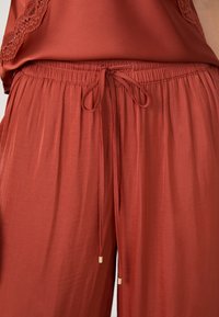 Roestkleurige loungebroeken van zacht, lichtgewicht materiaal. Voorzien van een elastische tailleband met een trekkoord en gouden hardware accenten.