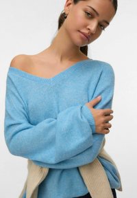 Pull oversize bleu clair avec un col en V et des manches larges. Texture douce, poignets côtelés. Détail notable : un pull beige noué à la taille.