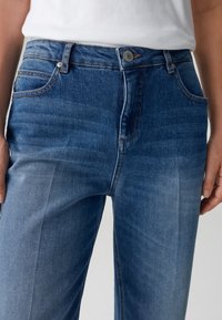 Jean en denim bleu avec une taille mi-haute, cinq poches et des coutures contrastées. Le tissu a une texture lisse et un léger effet délavé.