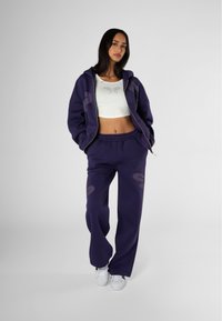 Felpa viola con ampi motivi grafici, abbinata a pantaloni della tuta larghi coordinati. Top corto bianco con dettagli in strass delicati. Sneakers bianche.