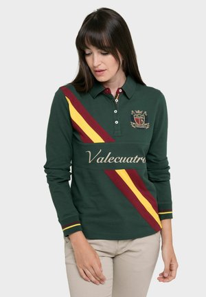 Camisa polo de manga larga verde con rayas diagonales en burdeos y amarillo, logo bordado y texto "Valecuatro" en el pecho.