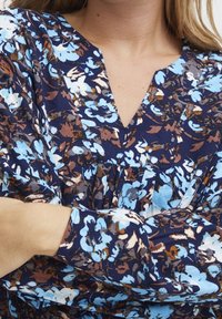 Blusa floreale in blu scuro con motivi blu e marroni. Presenta un design a scollo a V e maniche lunghe con un leggero dettaglio plissettato. Tessuto morbido.