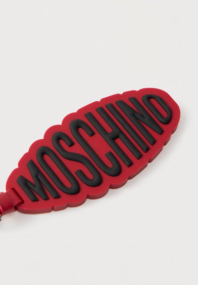 Κόκκινη λαστιχένια μπρελόκ σε σχήμα φύλλου με ανάγλυφα μαύρα γράμματα που γράφουν "MOSCHINO" σε λευκό φόντο.
