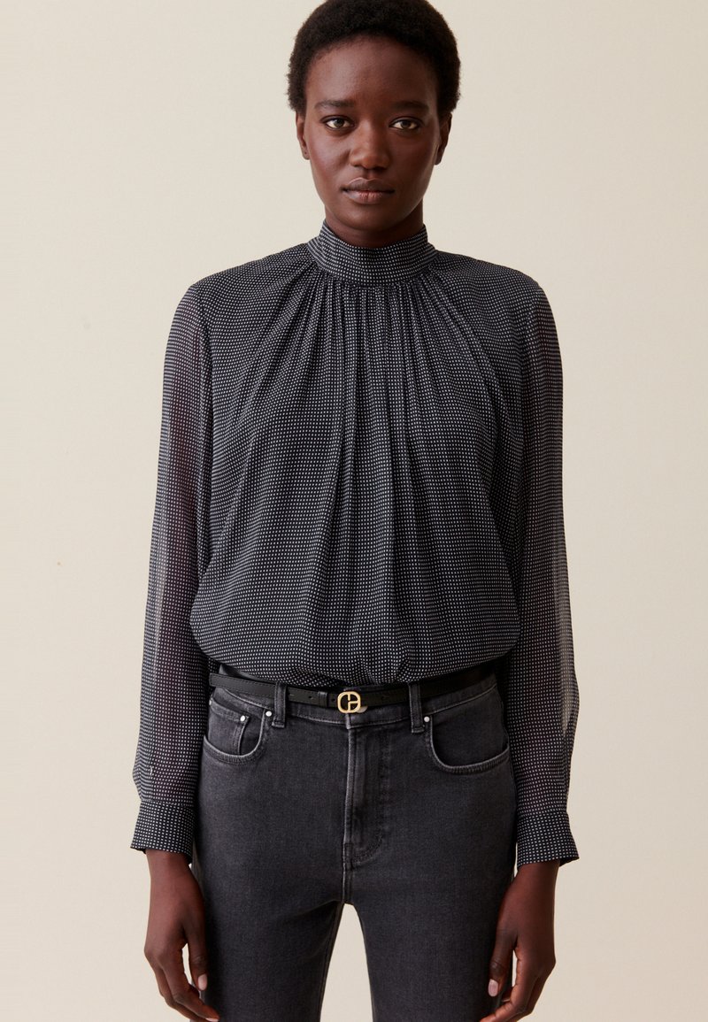 Mujer vistiendo una blusa negra transparente de manga larga y cuello alto con puntos blancos, metida en unos jeans gris oscuro con un cinturón negro.