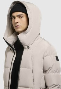 Beige Daunenjacke mit hohem Kragen und großer Kapuze, ausgestattet mit einem Frontreißverschluss, schwarzen Akzenten und einem gesteppten Design für mehr Struktur.