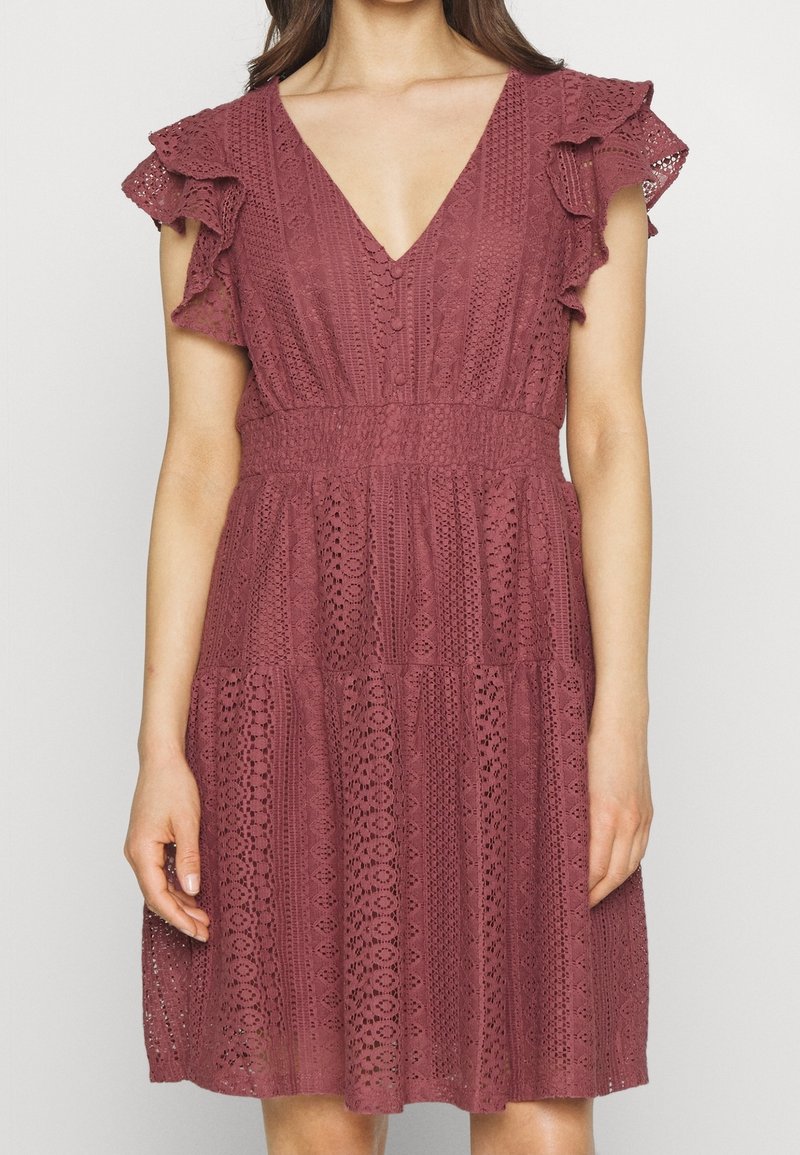 Femme portant une robe sans manches en dentelle rose poudré avec un décolleté en V et des détails à volants aux épaules.