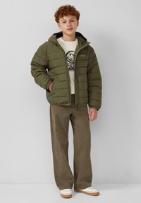 Olivgrüne, gepolsterte Jacke mit Kapuze, zwei seitlichen Taschen und einem Logo. Kombiniert mit einem beigen Rundhals-Sweatshirt und braunen weiten Hose.