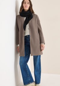 Abrigo marrón claro con un corte ajustado, suéter beige, jeans de pierna ancha azules y una bufanda negra. Tejido suave con detalles de botones.