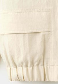 Gros plan d'un tissu beige avec une couture horizontale et un ourlet élastique froncé sur un vêtement.