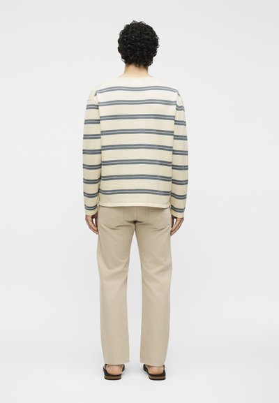 Personne aux cheveux bouclés foncés portant un haut à manches longues crème et gris à rayures, un pantalon beige et des sandales, debout tournée vers l'arrière.