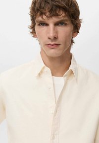 Camicia crema a bottoni realizzata in cotone, con colletto classico, maniche lunghe e una texture liscia. Due bottoni visibili sul davanti.